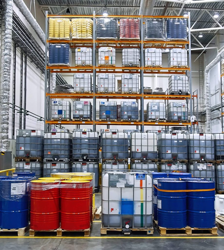 Ein Lagerraum mit gestapelten Fässern und Behältern in verschiedenen Farben auf Paletten.